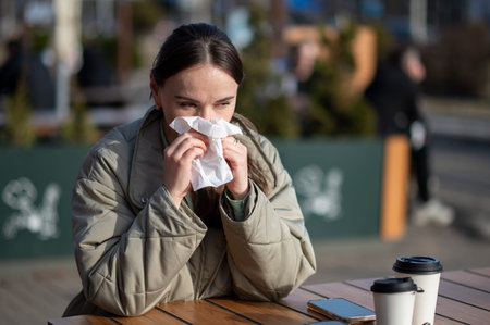 A concerned young woman in a jacket sits at a terrace table, holding a paper napkin to her face. Two...