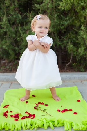 Cute little blond girl in a white dress posing on light green litter in the park  Summer の写真素材