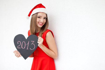 Beautiful young woman in a red Christmas hat and red dress, holding a sign in the form of a heart and the words  2013  over white wall background  A series of photos in my portfolio の写真素材