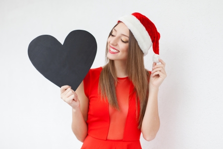 Beautiful young woman in a red Christmas hat and red dress, holding a sign in the form of a heart over white wall background  A series of photos in my portfolio の写真素材
