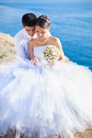 Couple in love young bride and groom dressed in white hugging on cliff background of blue sea in their wedding day in summerの写真素材