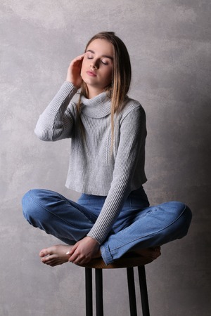 Sitting teen in pullover touching her face, girl with crossed legs, high fashion look, sitting girl, beautiful girl, blonde girl, isolated, model in studio, girl wearing jeans and sweater, long hair, gray backgroundの写真素材