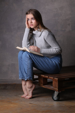 Sitting girl with book touching her head, high fashion look, sitting girl, beautiful girl, blonde girl, isolated, model in studio, girl wearing jeans and sweater, long hair, gray backgroundの写真素材