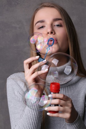 Girl in turtleneck blowing bubbles, bubble blower , high fashion look, beautiful girl, blonde girl, isolated, model in studio, long hair, close up, gray backgroundの写真素材