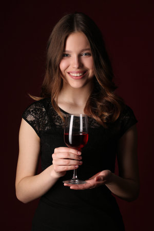 Standing lady with bocal, girl with wineglass, high fashion look, beautiful girl, brunette girl, isolated, model in studio, long hair, close up, dark red backgroundの写真素材