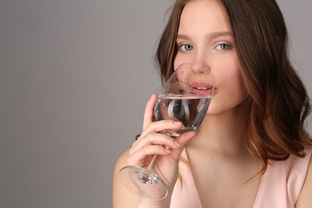 Girl drinking water from a wine glass, high fashion look, healthy lifestyle, beautiful girl, smiling girl, isolated, perfect make-up, big lips, model in studio, close up, gray backgroundの写真素材