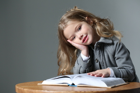 Little lady sitting at the table with a book, happy childhood, high fashion look, little girl, happy family, cute baby, beautiful girl, blonde girl, isolated, baby in studio, long hair, close up, gray backgroundの写真素材