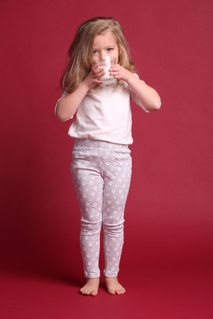 Kid in pajamas drinking milk, happy childhood, little girl, happy family, fashion clothes, cute baby, blonde girl, isolated, baby in studio, red backgroundの写真素材
