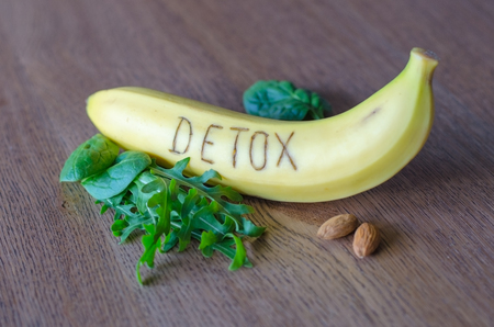 close-up of yellow banana with written word Detox, few rucola and spinach leaves and two almonds lying on wooden table backgroundの写真素材