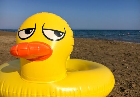 Yellow inflatable duck with frown annoyed, disgrunted expression on its face laying on the beach.の写真素材
