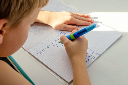 Kid hands doing math homework. Close up of young boy hand holding pen and writing a math homework close-up. concept of education at home.の写真素材