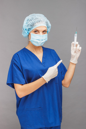 Woman doctor in the uniform, beret and sterile gloves with syringe in the hand, gray backgroundの写真素材