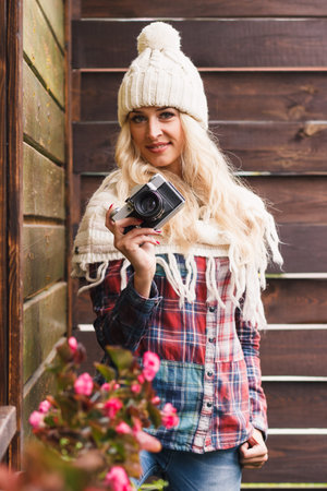 Young smiling blonde in the knitted hat holds a camera, soft focus background