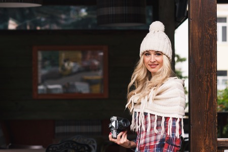 Young smiling blonde in the knitted hat holds a camera, soft focus backgroundの写真素材