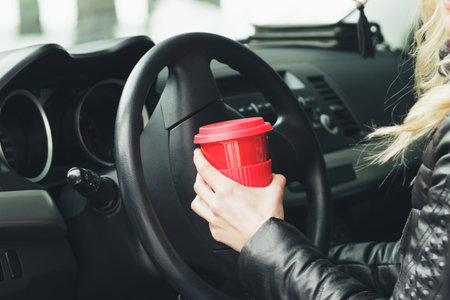 Woman with a red cup of hot drink keeps the wheel of a car, soft focus backgroundの写真素材