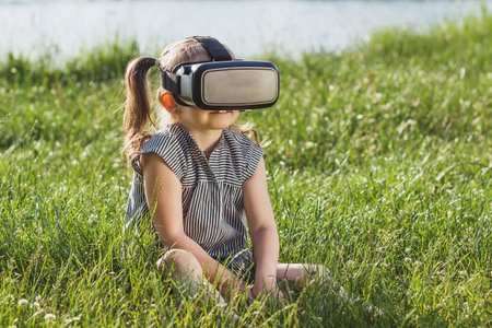 Little beautiful girl plays game with virtual reality glasses outdoors. Digital vr device
