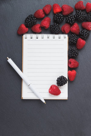 Red raspberries, blackberry, notebook and a pen on the black slate stone background, top viewの写真素材