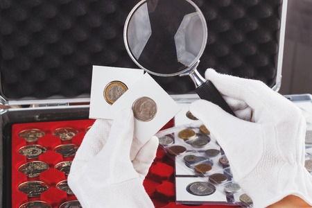 Hands in the gloves hold magnifying glass and gold collector coins in the flips, soft focus backgroundの写真素材