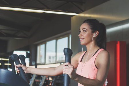 Sporty young woman makes sport exercises on the electrical trainer in the gym, soft focus background