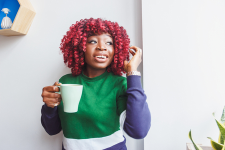 Dark-skinned woman dressed in casual clothes holding cup of hot drink, enjoying coffee or tea, soft focus backgroundの写真素材