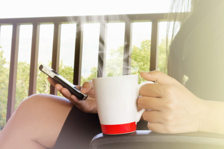 Women hand hold cup of hot drink while sitting and surfing the internet on mobile phoneの写真素材