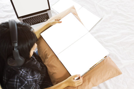 Asian girl sitting on bed reading blank book listening to music with headphone in relax mood with...