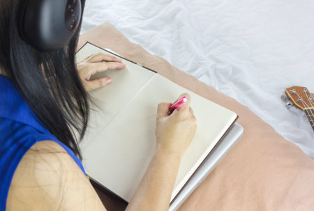 Women or girl sit on bed, listen to music and writing down on blank note book or making memo on empty memorandum and diary in relax mood with copy space, girl or women hand writing down on blank page on note book, homework or work on bed, idea on bedの写真素材
