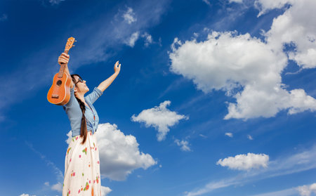 Asian women smiling happily holding ukulele and raise two arms embrace blue summer sky with puffy clouds, cheerful girl standing on blue sky background with freedom moodの写真素材