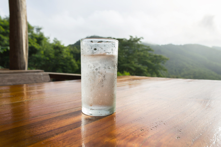 Single plain cool water and ice in glass on wood table with nature mountain view in the morningの写真素材