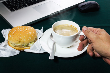 hand take hot coffee, burgerr and coffee on working table, easy meal with background of notebookの写真素材
