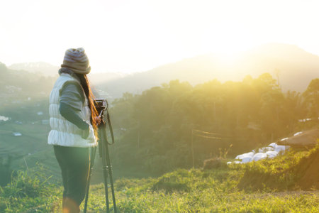 Back or rare view women shooting sunrise on mountain in green nature background, women wearing winter suit taking photo of sunrise in the morningの写真素材