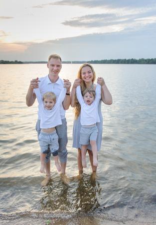 Happy family - father, mother, two sons on the beach with their feet in the water at sunsetの写真素材