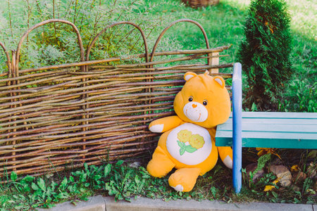 Adorable yellow stuffed big toy teddy bear on the ground near to a wicker fence and a benchの写真素材