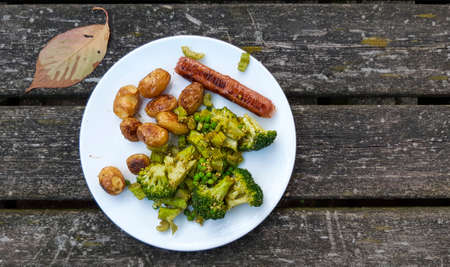 Vegan meal with oven baked potatos, plant based sausace, broccoli, peas and pesto on a white plate. Veganism and delicious food on weathered wooden tableの写真素材