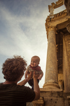Man with curly brown hair holding a baby in front of historical pillars from an old roman templeのeditorial素材