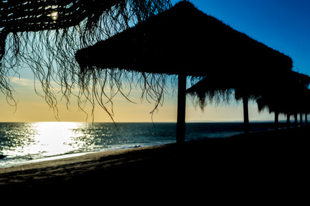 Serene vacation beach setting during a sunset. Silhouetted sunshades or thatched-roof huts and gradient of warm colors near the horizon, transitioning into cooler blue tones aboveの写真素材