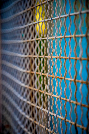 Close-up f a rusted expanded metal mesh featuring a repeating diamond pattern. The image showcases industrial texture and geometric rhythm, with a softly blurred blue and yellow background adding contrast and depthの写真素材
