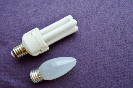 Two light bulbs. White energy-saving fluorescent bulb with four tubes, with a silver base and an oblong incandescent bulb with gray glass on a purple cloth background.の写真素材