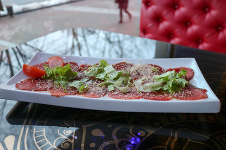 Meat appetizer of red beef and pork sprinkled with cheese and greens on a white rectangular plate on a table in a cafe, restaurant.の写真素材