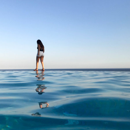 A beautiful slender girl in short shorts walks along the edge of the water of a luxurious infinity pool merging with the horizon on the background of the sea in a tropical warm sea resort.の写真素材