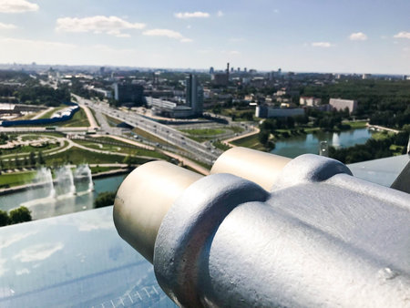 Viewing coin stationary binoculars against a white-blue sky in profile. Gray metal binoculars on the viewing platform at a height. From the roof overlooking the beautiful green city.の写真素材