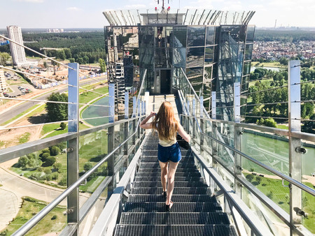 A beautiful girl, a woman with long hair in the hair comes down the metal stairs with a railing on the roof of a glass skyscraper, a tall building with a view of the big city from a height metropolis.の写真素材