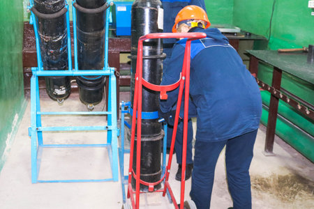 A worker in a helmet is loading a gas oxygen cylinder onto a trolley at an industrial plant.