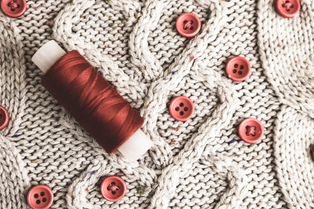 Beautiful texture of a soft warm natural sweater, fabrics with a knitted pattern of yarn and red small round buttons for sewing and a skein of red thread. The background.の写真素材