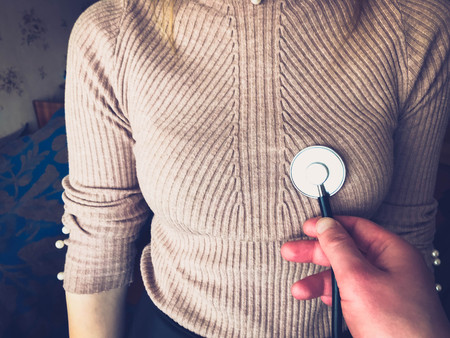 The doctor holds in his hand a medical stethoscope, a phonendoscope and listens to the chest, heart and lungs of a beautiful girl, a woman in a jacket, sweater.の写真素材
