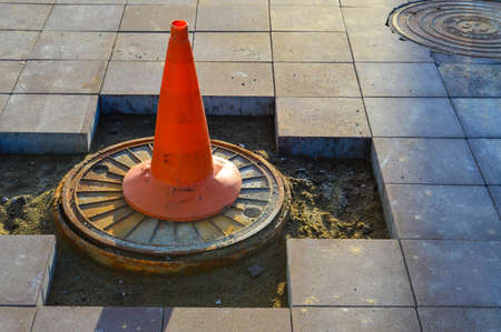 a bright orange cone stands on the sewer drain. marking of dangerous sections on the road, entry for people is prohibited. the cone stands on the broken asphalt, the builders are laying a new surface.の写真素材