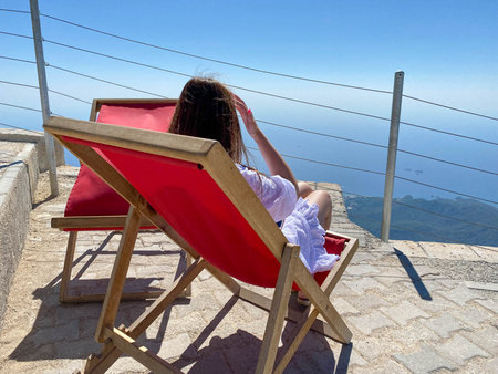 A woman sits on a sun lounger on a viewing platform high in the mountains overlooking the sea. Tourist rest in the mountains and at sea.の写真素材
