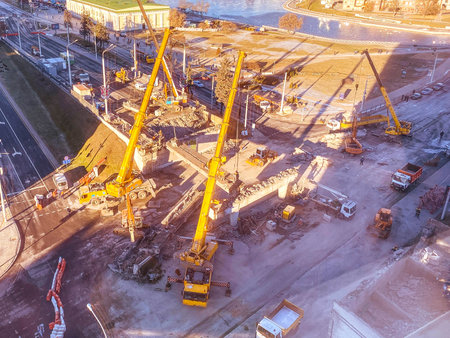 construction of a new overpass in the city center. near the river, walking path and many buildings. tall, yellow construction cranes are building a bridge from heavy beams.の写真素材