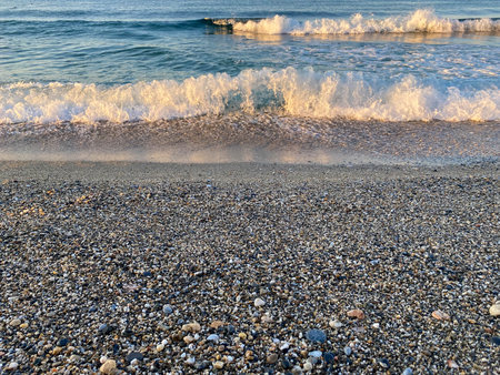 Waves of water in the sea and sand with small natural multi-colored stones on the seashore, small...