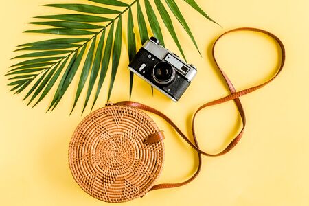 Traveler accessories concept on yellow background. Fashionable handmade natural round rattan bag, retro camera and tropical leaves.の写真素材